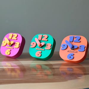 May include: Three colorful clocks with different colored hands and numbers. The clocks are made of a soft, textured material and have a playful, whimsical design. The clocks are labeled "ThicCloc" and have the numbers 12, 3, 6, and 9.