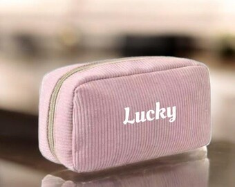 May include: A light pink corduroy cosmetic bag with a zipper closure. The word "Lucky" is printed in white on the front of the bag. The bag is rectangular and has a soft, textured appearance.