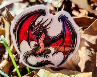 May include: A black dragon with red wings is depicted on a white sticker. The dragon is facing forward with its mouth open and its wings spread out. The sticker is on a background of fallen leaves.