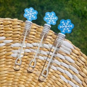 May include: Three clear bubble wands with white handles and snowflake-shaped toppers. The snowflake toppers are light blue with white snowflake designs. The wands are arranged on a woven surface with a natural and white pattern.