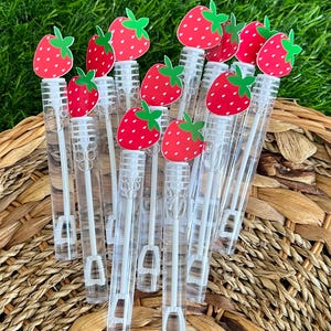 May include: Clear plastic bubble wands topped with red strawberry cutouts with green leaves. The wands are arranged in a group, with the strawberry tops visible. The background is green grass and a woven basket.
