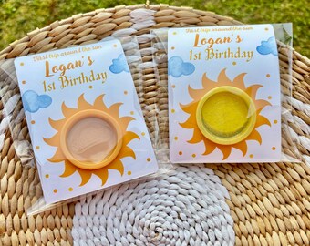 May include: Two party favors in clear plastic packaging. Each favor features a yellow sun design with the text "First trip around the sun" and "Logan's 1st Birthday". The favors are on a woven surface.