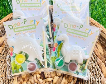 Puede incluir: Dos bolsas de regalo de plástico transparente que contienen una figura de dinosaurio de yeso blanco, un pequeño pincel de madera y dos pequeños botes de pintura redondos. La tarjeta adjunta dice Dino-Party, Alexander's 8th Birthday, con globos.