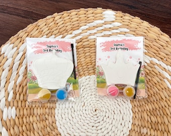 May include: Two paint-your-own crown kits for a birthday. Each kit includes a white crown, paint, and a brush. The packaging reads "Sophia's 3rd Birthday". The kits are on a woven surface.