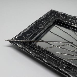 May include: A silver hair pin lies on a decorative, black framed mirror. The mirror has a reflective surface with geometric lines. The frame has a textured, ornate design with silver highlights.
