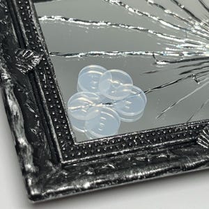 May include: A silver framed mirror with a cracked glass surface. The mirror is reflecting a cluster of clear, round, plastic buttons.