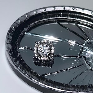 May include: A silver-toned stud earring with a large, sparkling, round, clear gemstone at its center. The earring is surrounded by smaller, clear gemstones. The earring is displayed on a cracked mirror surface within a dark, oval-shaped frame.
