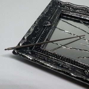 May include: A dark metal pen resting on a decorative, ornate mirror. The mirror has a black frame with silver accents. The mirror's surface is reflective, with a cracked glass design. The pen is thin and metallic.