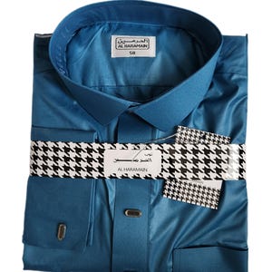 May include: Teal-colored button-down shirt with a classic collar and long sleeves. The shirt is neatly folded and packaged with a black and white houndstooth patterned band. The brand name "AL HARAMAIN" is visible on the label.