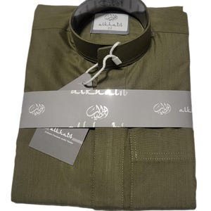 May include: Olive green long-sleeved shirt with a mandarin collar, neatly folded and packaged. The shirt is wrapped with a gray band featuring Arabic script and the brand name "alkhatib". A tag with the brand name and "Exclusive Premium Arab Thobes" is attached.
