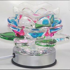 Puede incluir: Un difusor de aceites esenciales decorativo de vidrio con un diseño en forma de flor. Los pétalos de vidrio transparente están acentuados con tonos rosas, azules y verdes. La base es de color plateado con un borde negro y un cable de alimentación blanco.