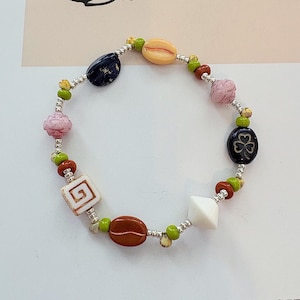 May include: A beaded bracelet featuring a variety of colorful beads. The bracelet includes beads in shades of pink, green, brown, white, and blue. The beads are strung together with silver-colored metal links.