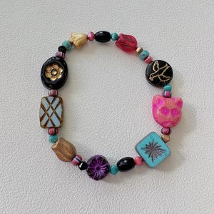 Op de afbeelding: Een kralenarmband met een verscheidenheid aan kleurrijke kralen. De armband bevat zwarte, roze, gouden, turquoise en paarse kralen in verschillende vormen, waaronder een kat, bloem en geometrische ontwerpen. De armband is geregen op een elastisch koord.