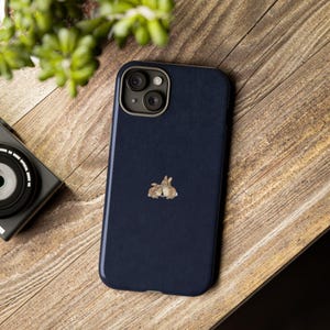 May include: A dark navy blue smartphone case with a textured finish featuring a small, centered illustration of two brown rabbits sitting together. The case has a rectangular camera cutout at the top left with two circular lenses visible.