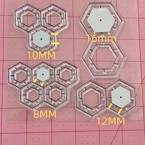May include: A set of four clear plastic hexagon-shaped stencils with different sizes. The stencils are on a pink cutting mat with white grid lines. The stencils are labelled with the following measurements: 10mm, 8mm, 12mm, and 16mm.