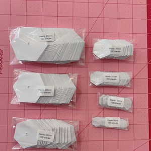 Puede incluir: Varias bolsas de plástico transparente que contienen pilas de piezas de papel hexagonales blancas. Cada bolsa tiene una etiqueta con "Hexie" y un tamaño en mm, junto con "100 pieces". El fondo es una esterilla de corte rosa.