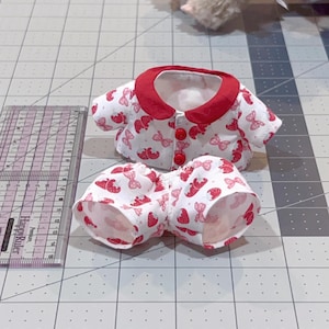 May include: A doll-sized pajama set featuring a white fabric with a red strawberry and bow pattern. The top has a red collar and three red buttons. The matching bottoms are also white with the same strawberry and bow pattern. A ruler is next to the pajamas.