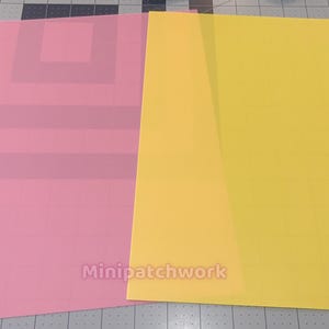 以下が含まれることがあります： ピンクと黄色の2枚の紙が、灰色のカッティングマットの上に置かれています。ピンクの紙には正方形のデザインの半透明のオーバーレイがあります。黄色の紙は無地です。「Minipatchwork」の文字がピンクの紙に表示されています。