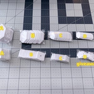 以下が含まれることがあります： A、B、C、Dとラベルされた白いタグのいくつかのパッケージが、グリッド状の表面に配置されています。 タグはさまざまな形とサイズで、ひもを通すための穴があります。 「giveaway」という単語は黄色で書かれています。
