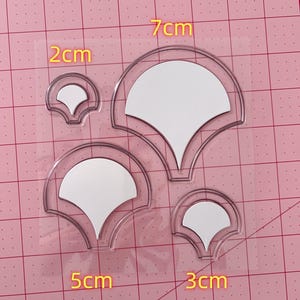 May include: Four clear plastic stencils in the shape of a scallop shell. The stencils are layered on top of each other and measure 2 cm, 3 cm, 5 cm, and 7 cm in diameter.