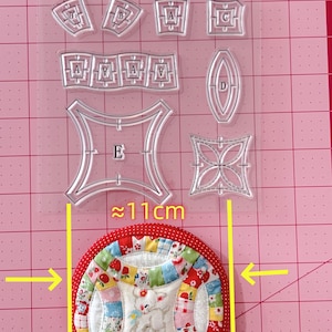 Może przedstawiać: Blok patchworkowy z tkaniny w kolorze czerwonym, białym i niebieskim z białym jagnięciem w środku. Blok ma około 11 cm średnicy i jest otoczony przez przezroczysty plastikowy szablon z literami i kształtami.