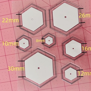 Puede incluir: Un conjunto de seis plantillas hexagonales de plástico transparente de diferentes tamaños, que van de 8 mm a 30 mm. Las plantillas están sobre una superficie cuadriculada rosa.