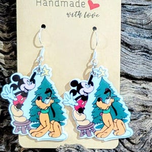 May include: A pair of dangle earrings featuring Mickey Mouse and Pluto in front of a Christmas tree. The earrings are attached to a tan card with the text "Handmade with love".