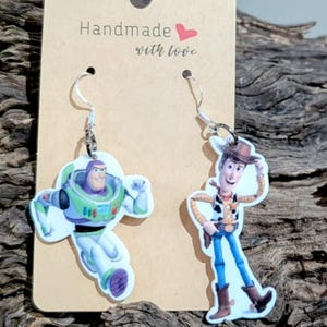Può includere: Orecchini fatti a mano con Buzz Lightyear e Woody di Toy Story. Gli orecchini sono attaccati a una carta marrone con la scritta "Handmade with love". Gli orecchini sono esposti su un pezzo di legno alla deriva.