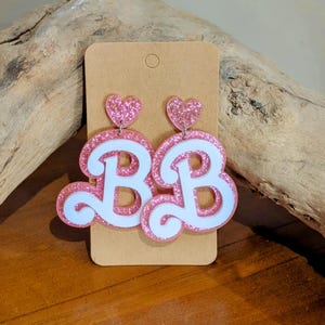 Puede incluir: Pendientes de acrílico rosa brillante con la forma de las letras "BB" con detalles blancos. Cada pendiente tiene un corazón rosa brillante en la parte superior. Se muestran en una tarjeta marrón.
