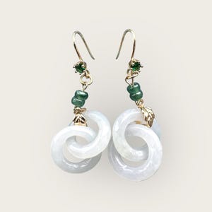 May include: Elegant dangle earrings featuring interlocking white jade rings. The earrings have gold-colored hooks, with green gemstone accents and small gold leaf details. A sophisticated accessory.