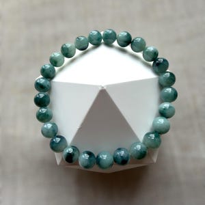 May include: A bracelet with teal and green polished beads. The beads are round and strung together to form a complete circle. The bracelet is displayed on a white geometric shape against a neutral background.