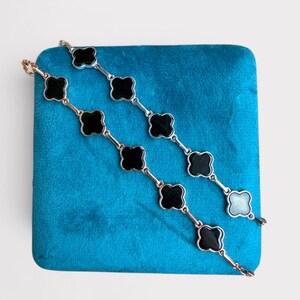Op de afbeelding: Twee armbanden met zwarte klavervormige bedels worden getoond op een blauw fluwelen oppervlak. Eén armband heeft goudkleurige schakels en de andere heeft zilverkleurige schakels. De bedels zijn zwart met een glanzende afwerking.