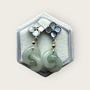 May include: A pair of elegant earrings featuring a four-petal flower design with a small gemstone center. The earrings have a silver-tone metal and are connected to a light green jade knot. The earrings are displayed on a gray velvet surface.