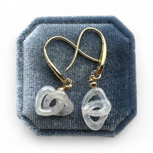May include: A pair of gold-colored dangle earrings with a unique design. Each earring features a gold hook and a pendant made of interlocking, translucent, light gray shapes. The earrings are displayed on a blue velvet surface.