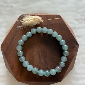 May include: A light blue beaded bracelet with round, translucent beads. The bracelet is displayed on a dark brown, wooden, hexagonal stand. A small, dried, beige floral accent is placed on the stand.