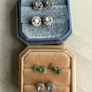 Puede incluir: Dos joyeros de terciopelo, uno azul y otro beige, cada uno con dos pares de pendientes. Los pendientes presentan piedras preciosas de color verde claro y engastes de metal plateado. Un juego tiene un diseño floral, el otro un diseño de lazo.