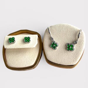 Puede incluir: Un conjunto de joyas con un par de pendientes de botón y un par de pendientes colgantes. Ambos conjuntos tienen un diseño de trébol con piedras preciosas verdes y metal plateado. Los pendientes se exhiben sobre una superficie beige dentro de una caja con ribete dorado.