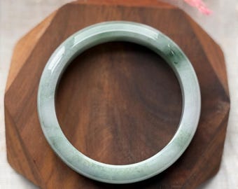 May include: A jade bangle bracelet with a pale green hue. The smooth, circular bracelet is displayed on a dark wooden surface. The jade has subtle variations in color, adding depth to the design. A pink decorative element is in the background.