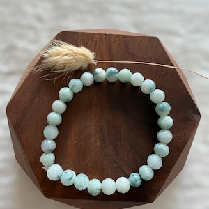 May include: A bracelet made of round, pale green beads with blue-green veining. The bracelet sits on a dark wooden hexagonal stand, with a dried floral decoration.