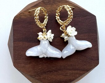 May include: A pair of gold-toned earrings featuring a whale tail design. The earrings have a gold wreath-like hoop, white flower accents, and small pearl-like beads. The whale tails are white and have a pearlescent finish. The earrings are displayed on a wooden surface.