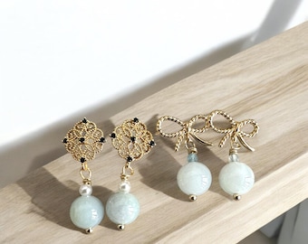 May include: A pair of gold earrings with a delicate filigree design and black accents. Each earring features a round, light blue gemstone and a small pearl. The earrings are displayed on a wooden surface.