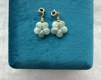 May include: A pair of flower-shaped earrings with a light green hue. The earrings are made of a translucent material and are accented with gold-colored metal. They are displayed on a teal velvet surface.