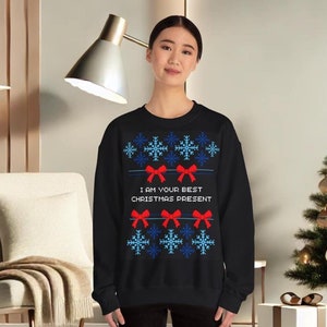 Könnte beinhalten: Schwarzer Weihnachtspullover mit einem blauen und weißen Schneeflockenmuster und roten Schleifen. Der Pullover hat den Text "Ich bin dein bestes Weihnachtsgeschenk" aufgedruckt.