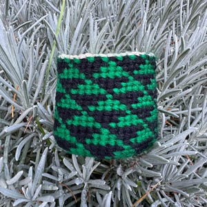 May include: A small, handwoven basket with a geometric pattern in black and green. The basket's rim is finished with a white border. The basket is set against a backdrop of silvery-green foliage.