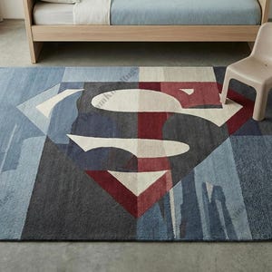 Pode incluir: Um tapete retangular com o logotipo do Superman no centro. O logotipo é branco, vermelho e azul, sobre um fundo de formas geométricas azuis, cinzentas e cinza escuro. Uma pequena cadeira bege está por perto.