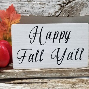 May include: White wooden sign with black cursive lettering that reads "Happy Fall Y'all". A small orange and red artificial pumpkin sits in front of the sign.