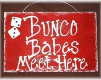 Może przedstawiać: Czerwony drewniany znak z białym napisem "Bunco Babes Meet Here". W lewym górnym rogu namalowane są dwie białe kostki do gry z czarnymi kropkami.