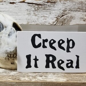 May include: White wooden sign with black text that reads "Creep It Real". The sign is sitting on a wooden surface next to a white skull with cobwebs.