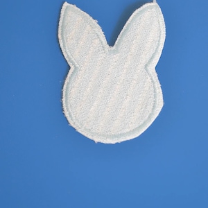 Peut inclure: Un écusson en forme de lapin bleu clair avec un aspect texturé semblable à de l'éponge. Le patch a un contour bleu clair et se détache sur un fond bleu uni. Idéal pour les créations sur le thème de Pâques ou du printemps.