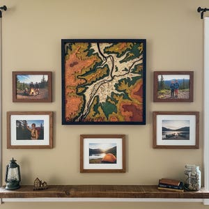 May include: A wall display featuring a large, framed topographical map in shades of green, orange, and brown. Surrounding the map are several framed photographs with outdoor themes, including camping and scenic landscapes. A rustic shelf below holds a lantern and decorative items.